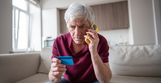 A concerned senior man holding a credit card and talking on a yellow smartphone, likely discussing a financial issue or seeking customer service. He appears puzzled and worried while on the phone.